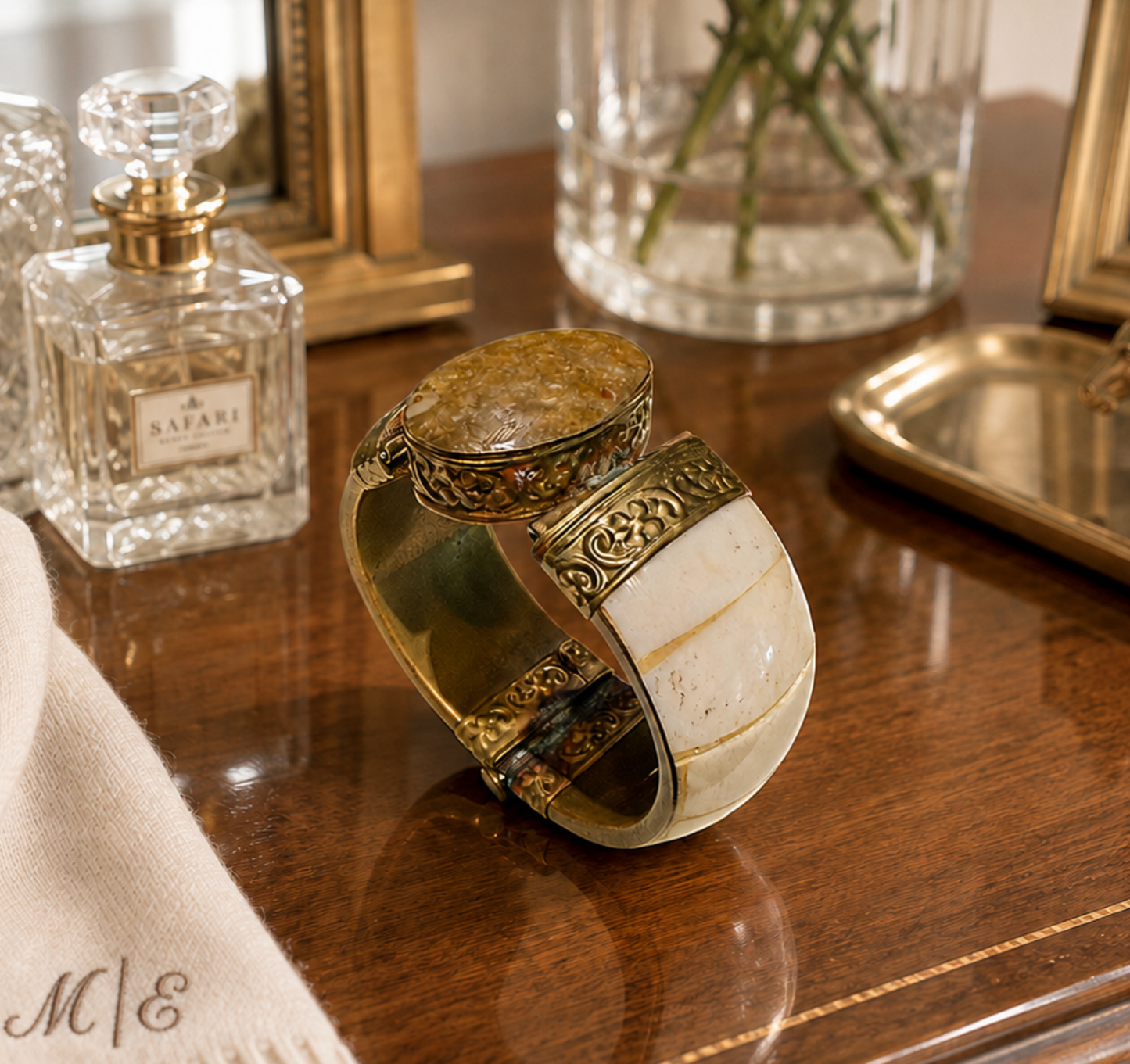 An antique-style cuff bracelet with a large oval cream-colored stone, gold-toned metal bands, and intricate filigree detailing, displayed on a polished wooden table next to a perfume bottle.