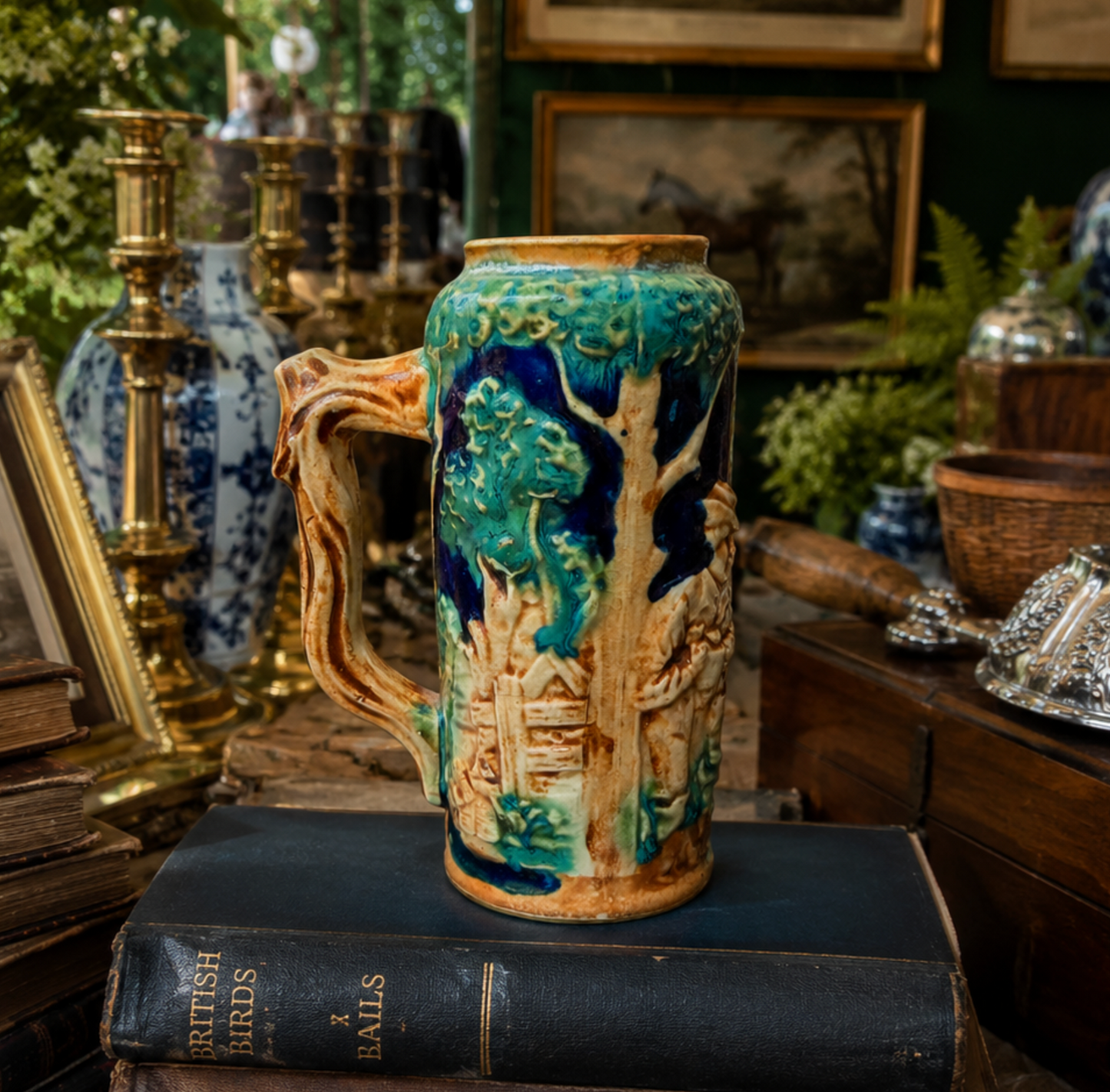 A vintage ceramic beer mug with a raised tree design, featuring turquoise, dark blue, and tan glazes, displayed on a dark book in a cluttered antique shop setting.