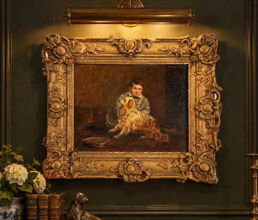 An ornate gold-framed classical painting hanging on a dark green wall, depicting a young boy holding a golden dog. The scene includes a brass wall sconce above, a vase of flowers and books below, and a small dog statue in the foreground.