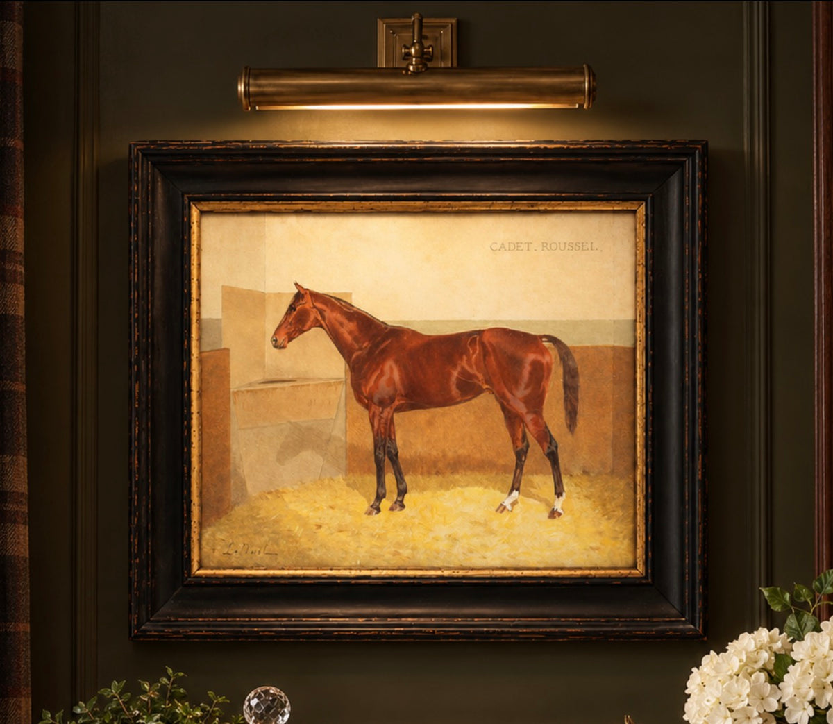 A framed portrait painting of the horse Galley Ranel, displayed on a dark green wall under a brass wall sconce. The painting features a brown horse standing in a stable with a beige background, enclosed in a dark wood frame.