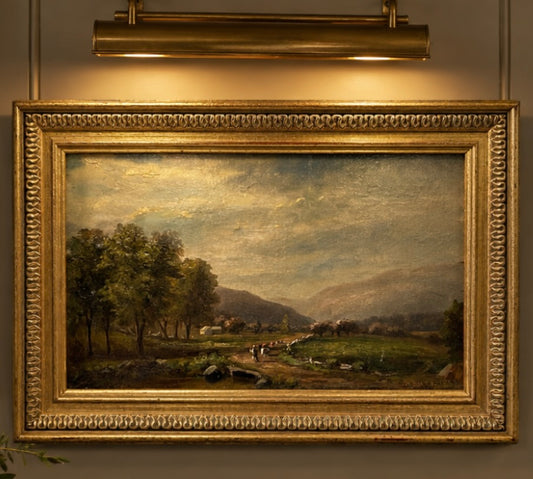 A framed landscape oil painting in a decorative gold frame, hanging under a brass wall sconce. The painting depicts a scenic valley with trees, a path, distant hills, and a cloudy sky.