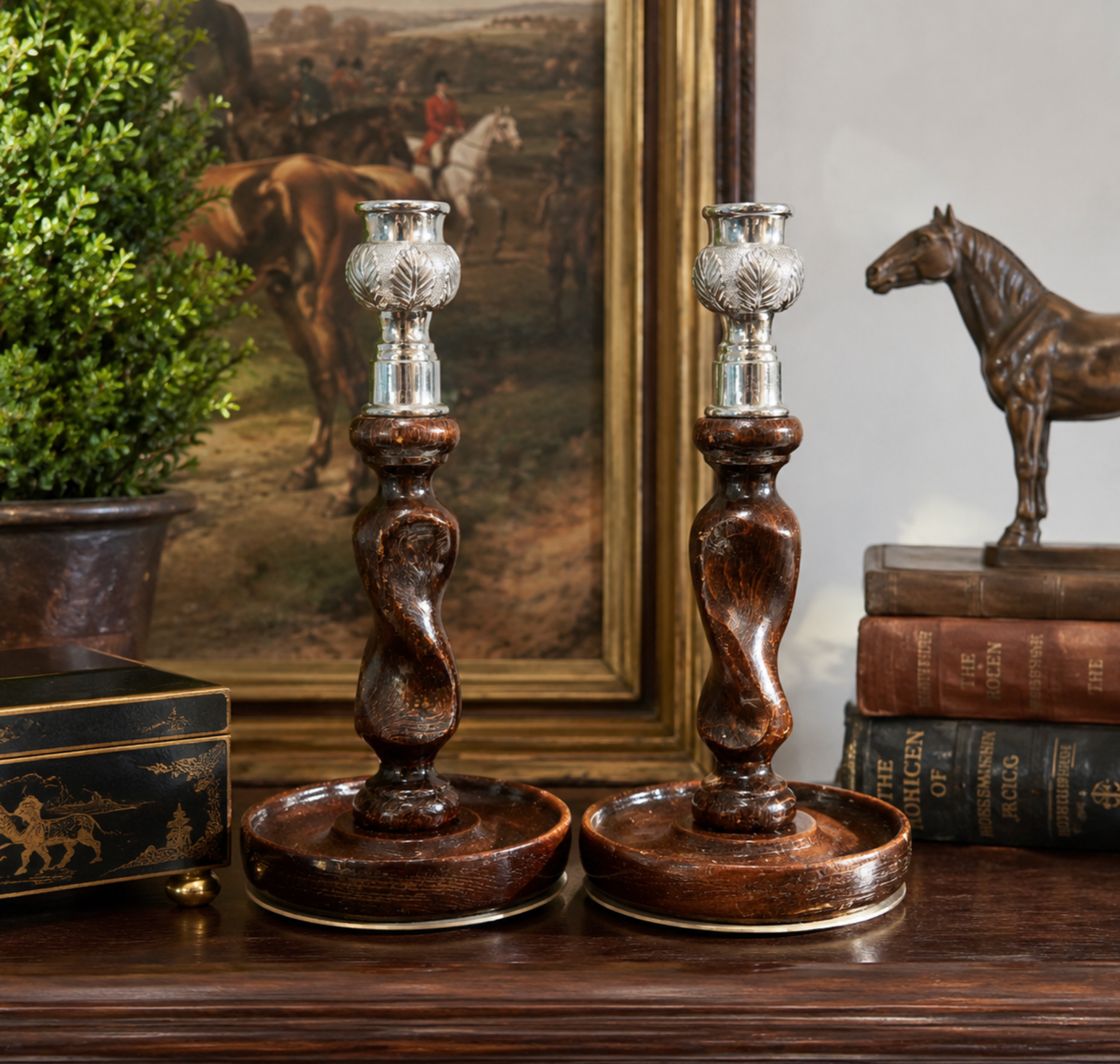 Two ornate dark brown wooden candlestick holders with twisted stems and silver-toned cup holders, displayed on a wooden table in a styled interior setting with books, a framed painting, and a small horse statue.