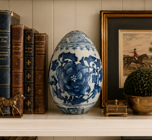 A large decorative ceramic egg-shaped vase with blue floral patterns on a white base, displayed on a shelf next to antique books, a framed equestrian painting, and a small gold-toned box.