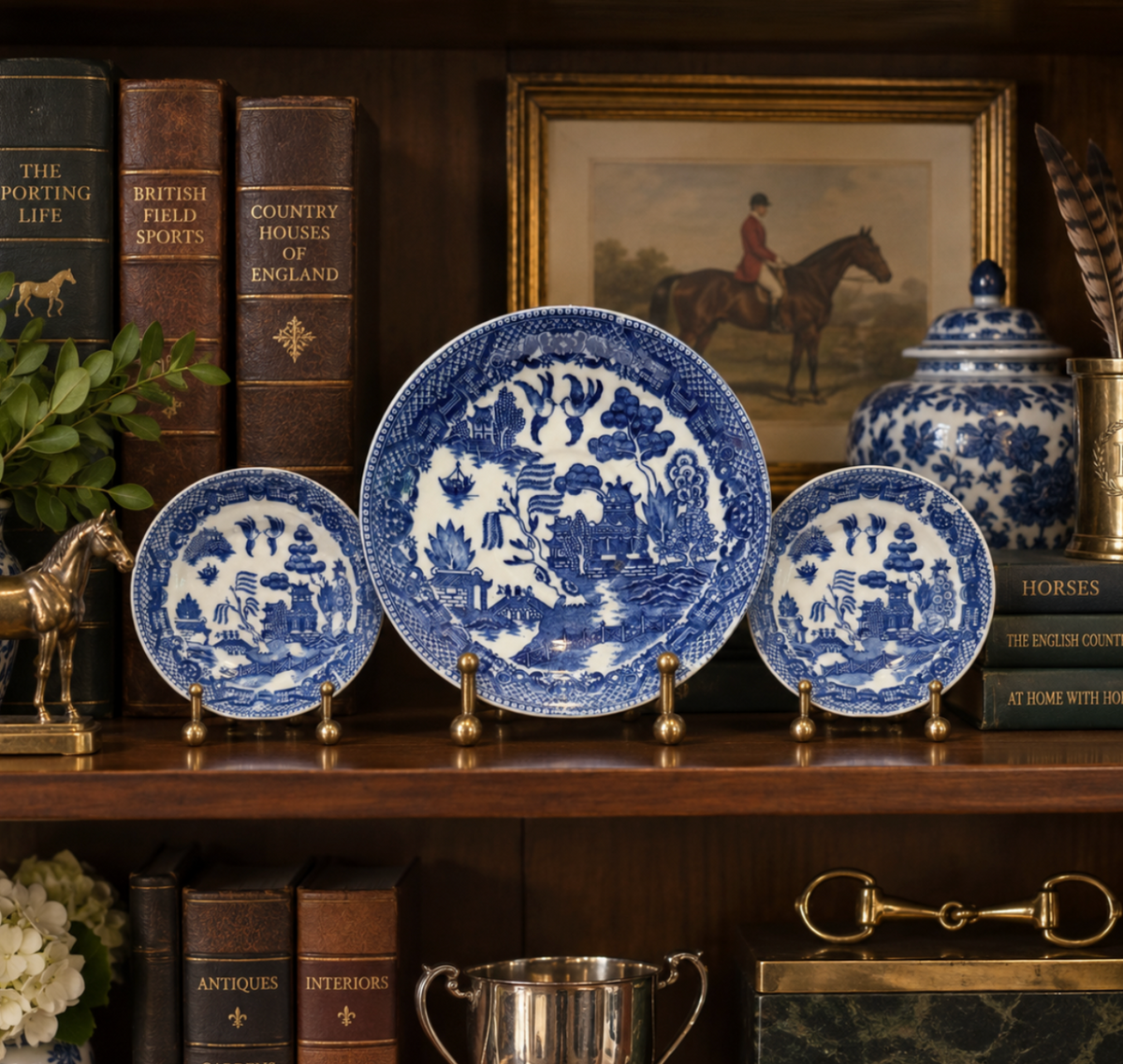 A set of blue and white ceramic plates and bowls displayed on a wooden shelf alongside vintage books, a framed equestrian painting, and decorative accessories.