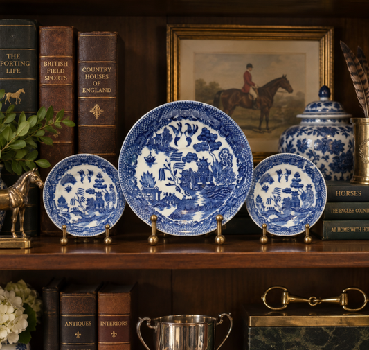 A set of blue and white ceramic plates and bowls displayed on a wooden shelf alongside vintage books, a framed equestrian painting, and decorative accessories.