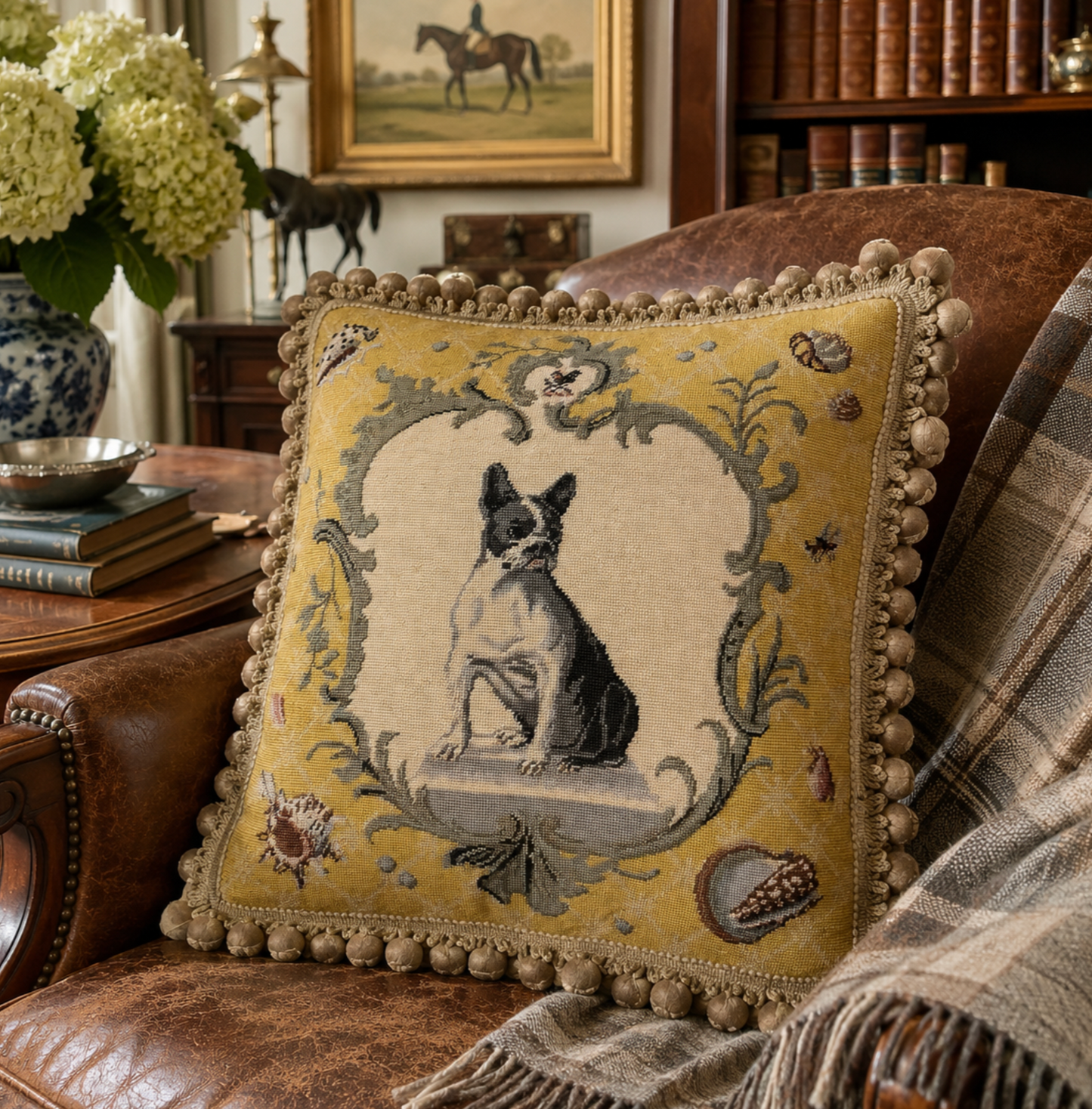A square decorative throw pillow with a pom-pom trim and a central embroidered dog motif, styled on a brown leather armchair in a cozy living room setting.