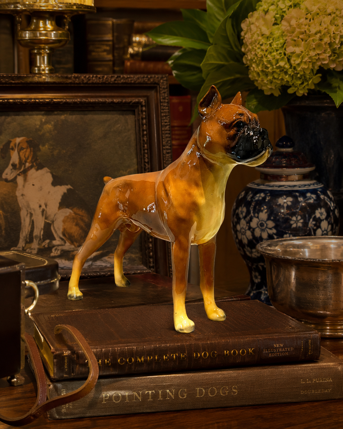A glossy ceramic figurine of a Boxer dog standing on a vintage book, styled in a rustic, antique-filled room with a blue-and-white patterned vase and framed dog paintings in the background.