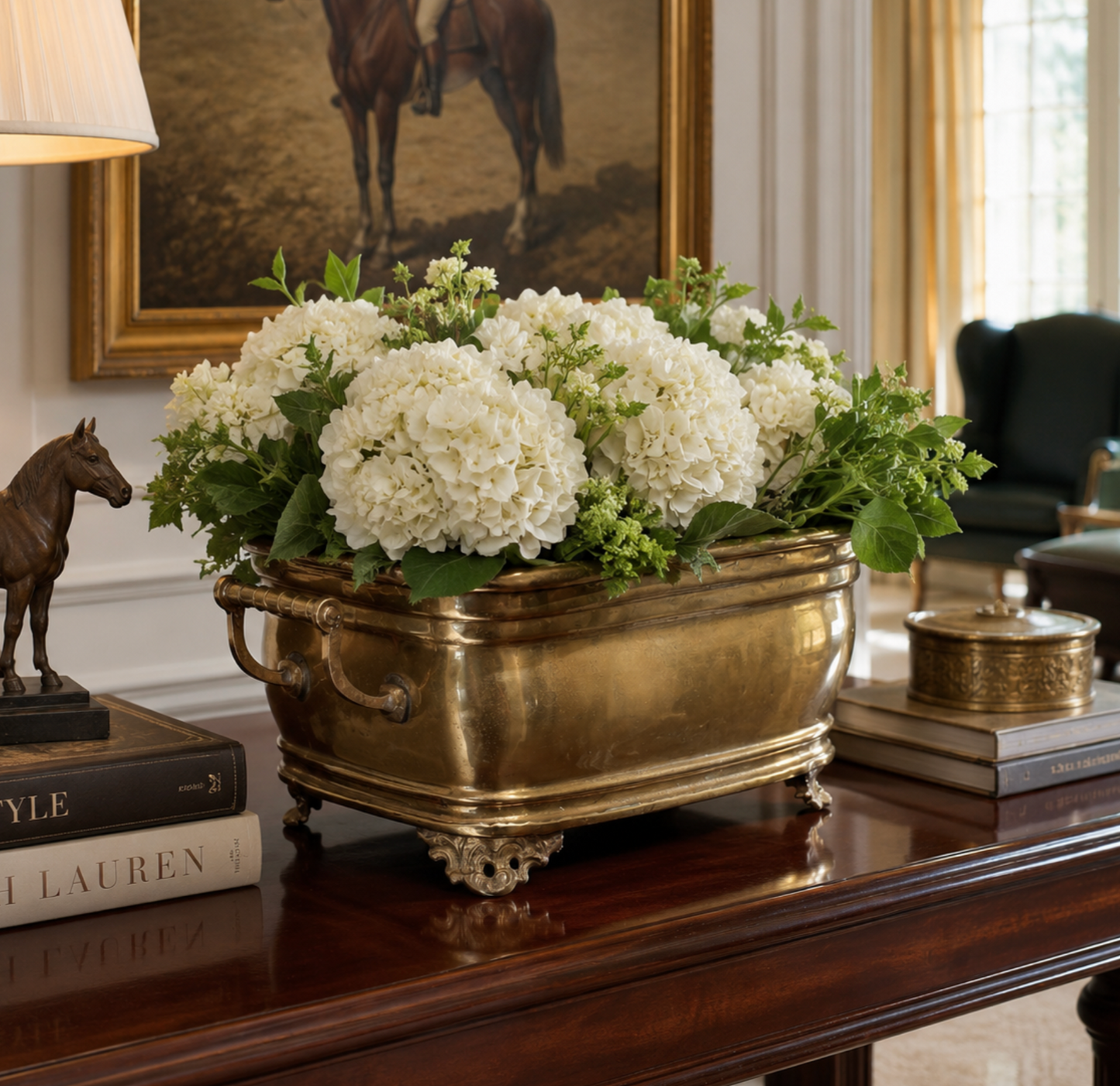 A large rectangular brass planter filled with white hydrangea blooms and green foliage, displayed on a polished dark wood console table in an elegant room setting.