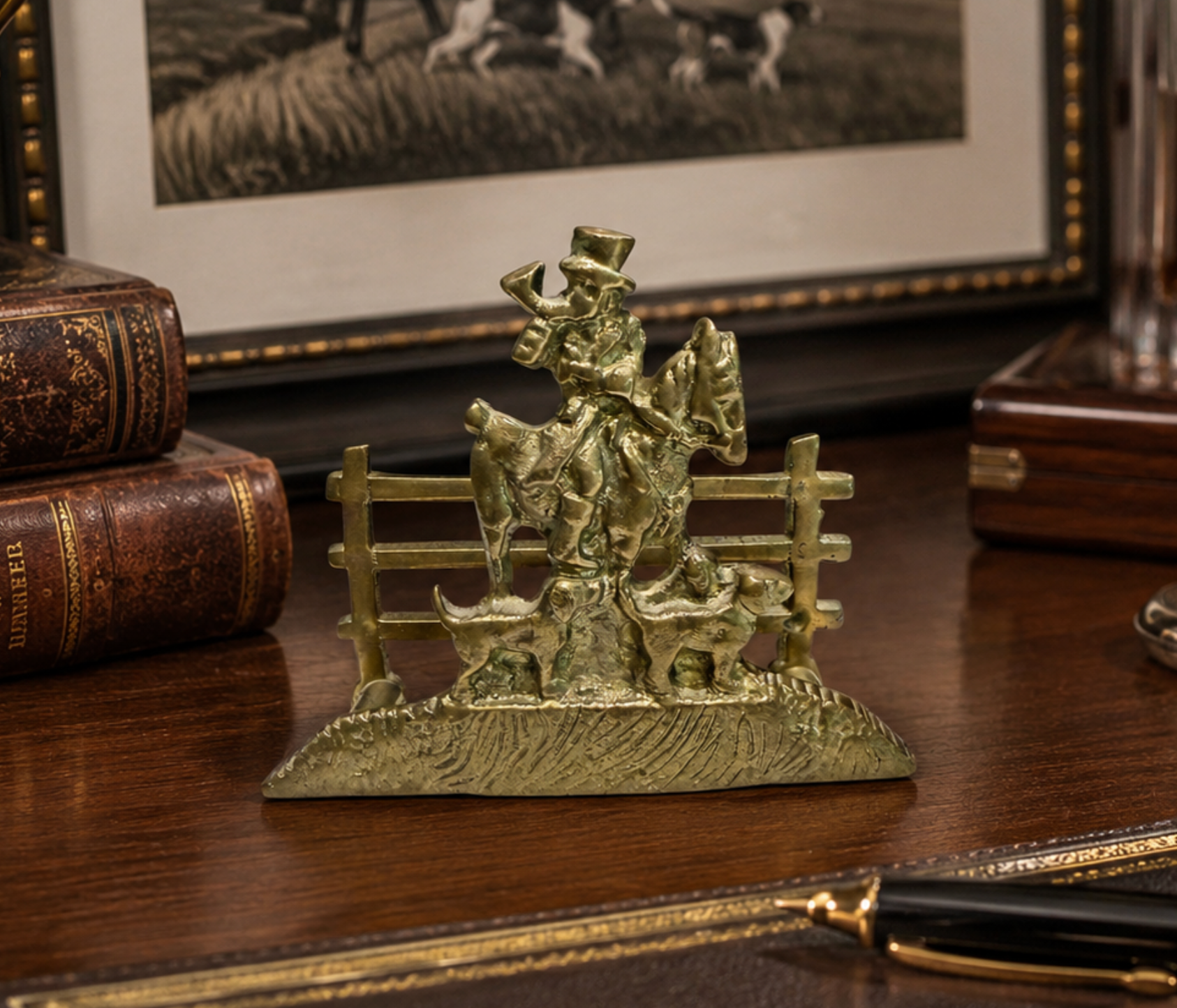 A small vintage-style bronze figurine of a cowboy riding a horse over a fence, displayed on a wooden desk with antique books and framed artwork in the background.