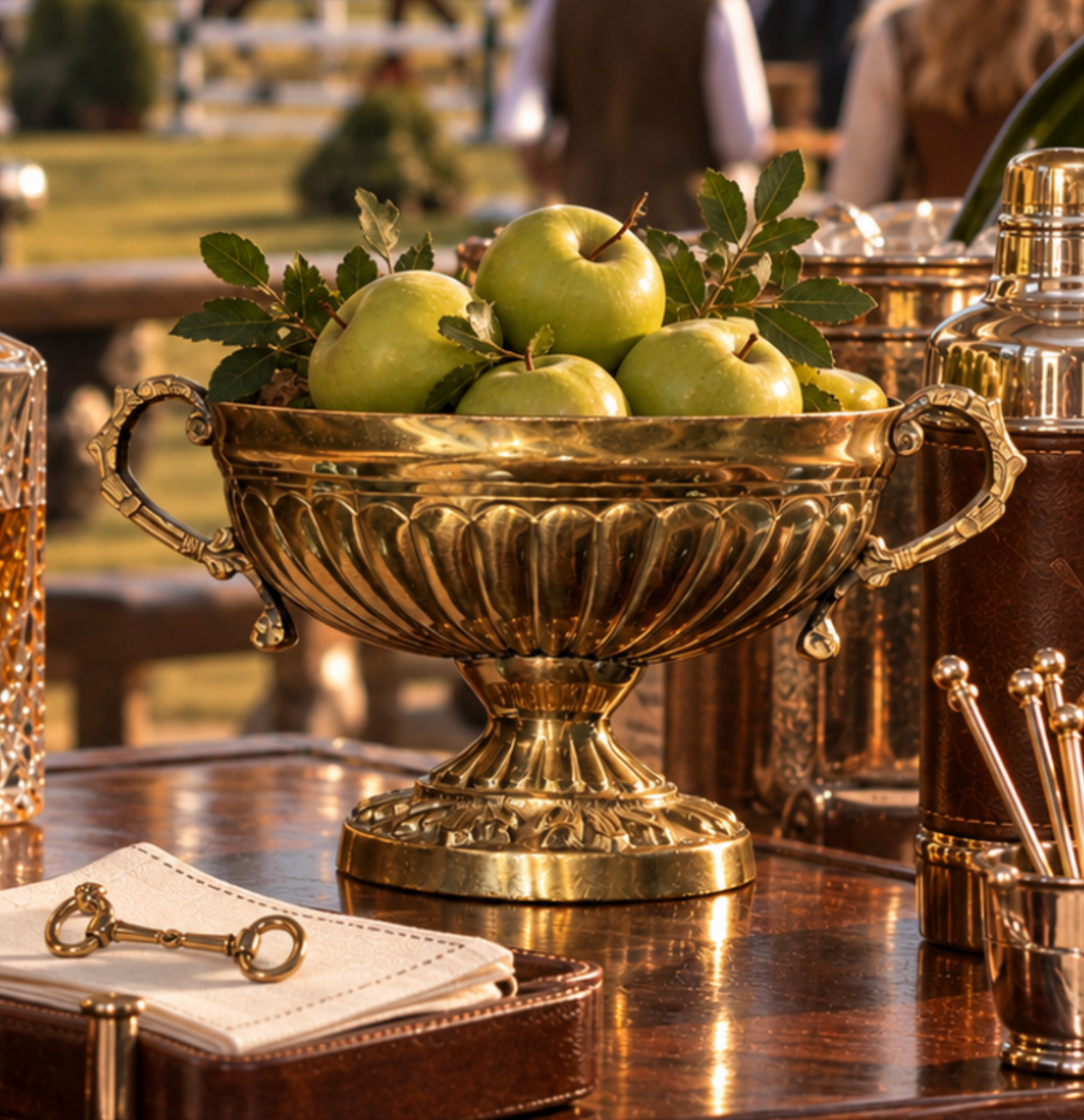 A decorative brass pedestal bowl filled with green apples and leafy stems, displayed on a polished wooden table in an outdoor event setting with barware and warm sunlight.