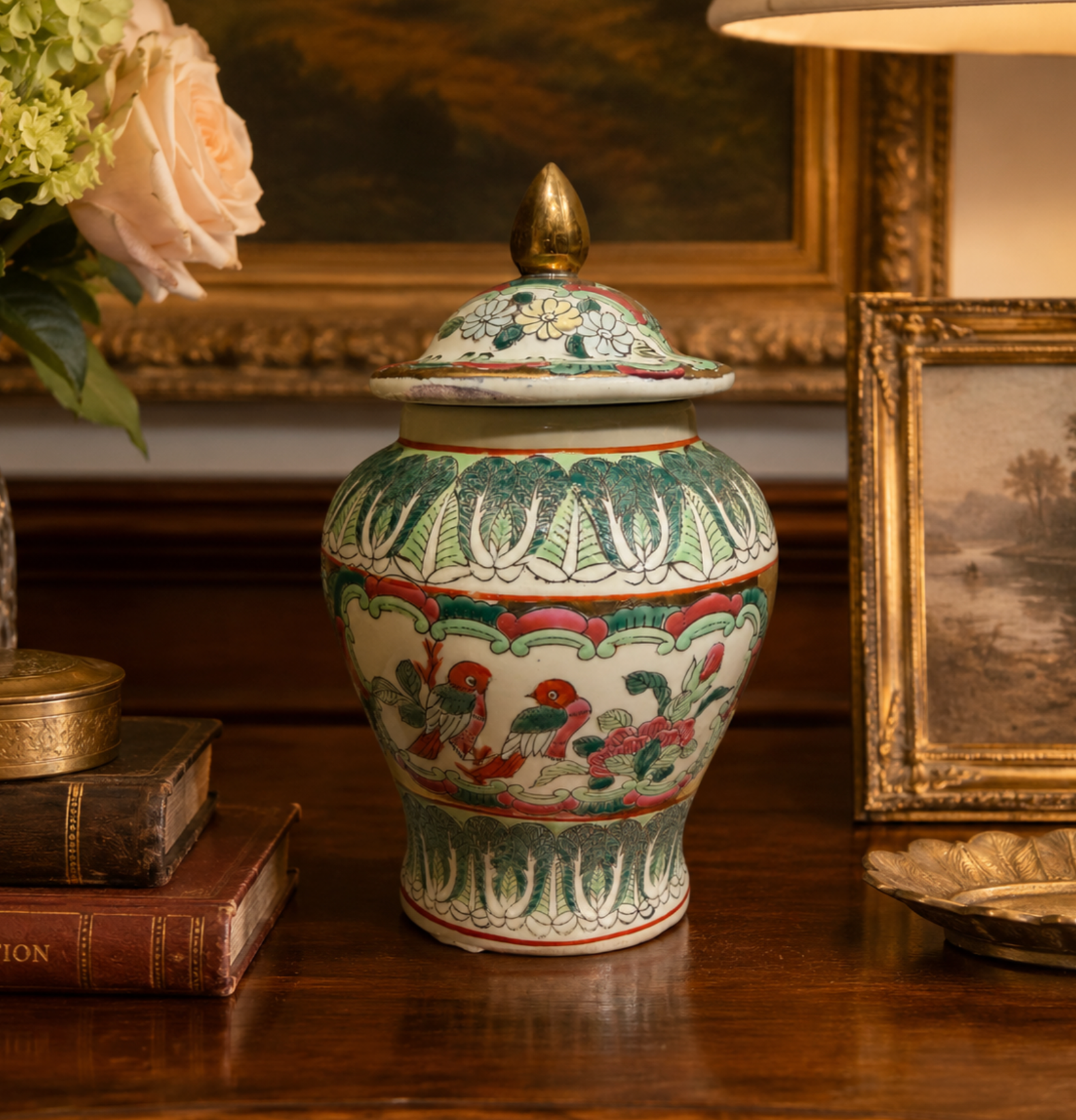 A vintage Chinese-style ceramic lidded jar with hand-painted floral and bird motifs in green, red, orange, and white, displayed on a wooden table in a styled room setting.