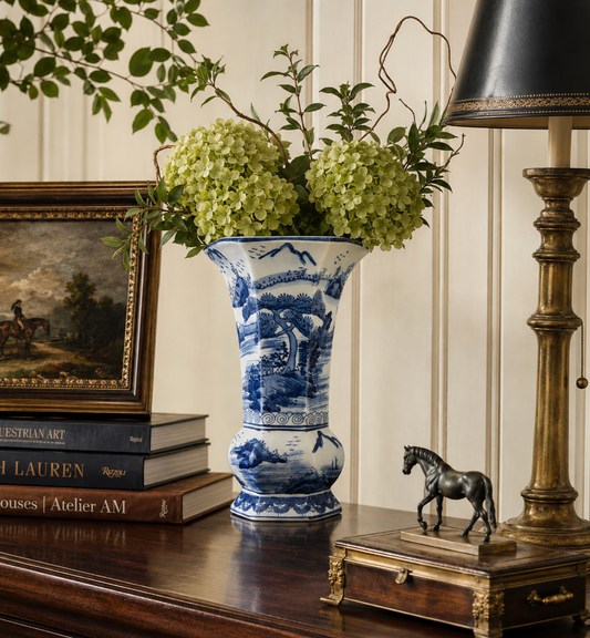A blue and white porcelain vase with a scenic landscape design, filled with green hydrangea blooms and placed on a dark wooden console table next to a brass lamp, books, and a small horse figurine.