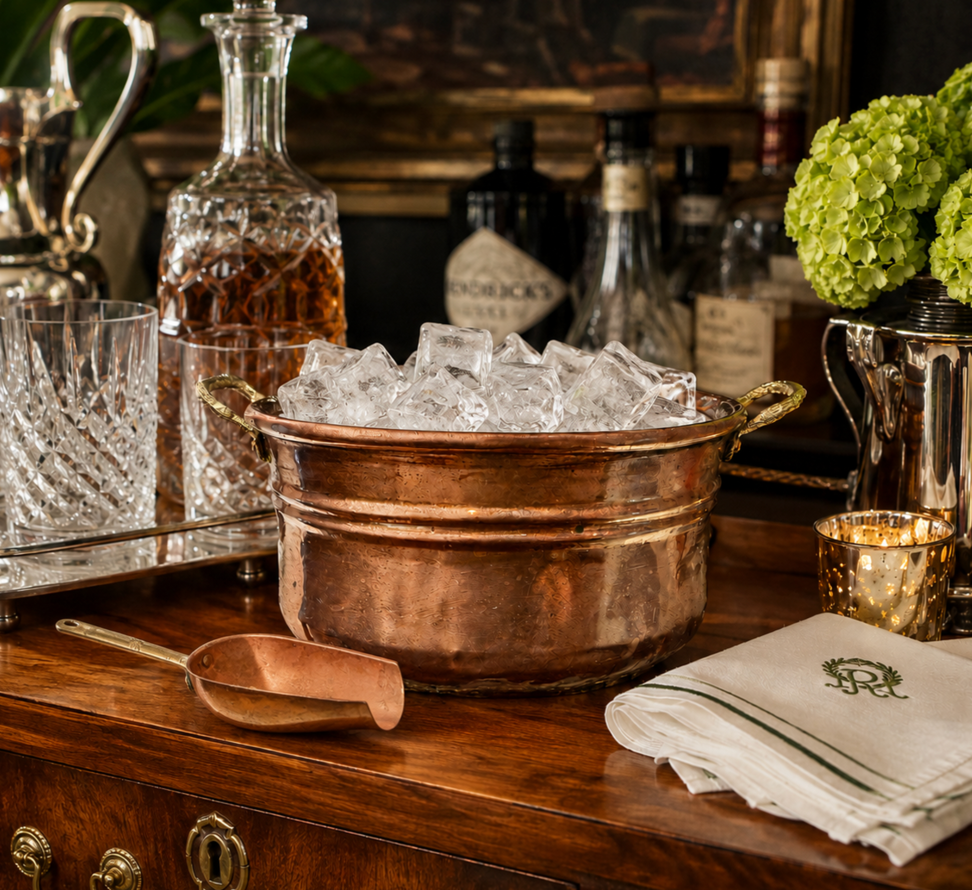 A copper double bucket ice bucket filled with ice, styled on a wooden bar counter with crystal glasses, a decanter, bottles, a scoop, folded white napkins with a green monogram, and green hydrangeas in the background.