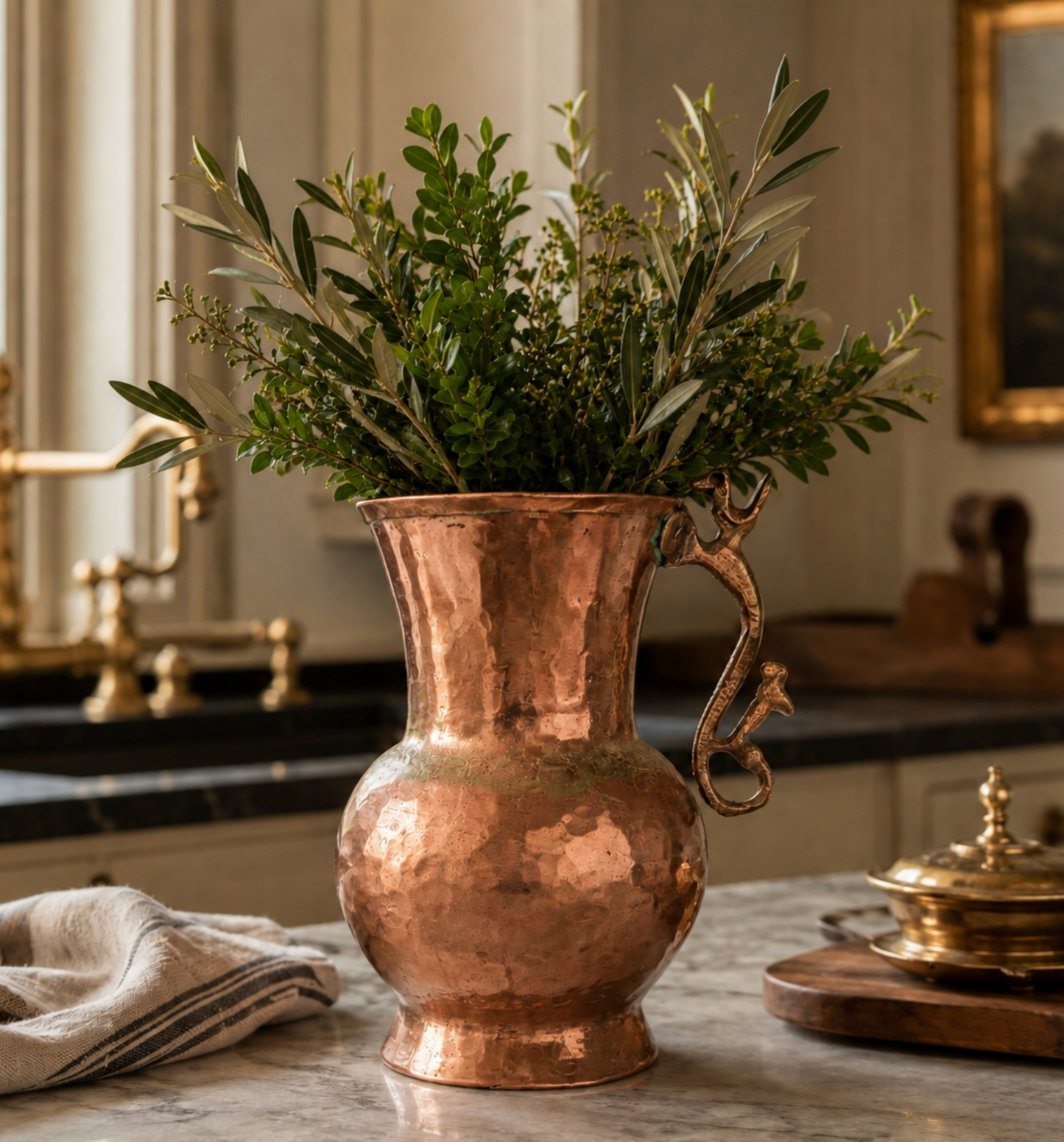 A hammered copper pitcher-style vase with a sculpted dragon handle, filled with green olive branches and foliage, set on a marble countertop in a warm, elegant kitchen.