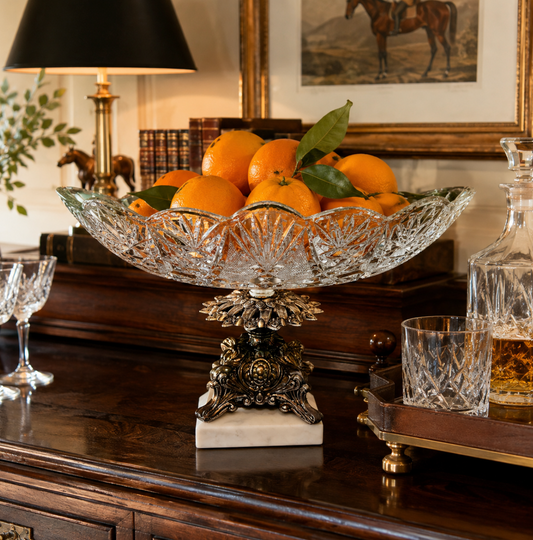 A clear cut-glass fruit bowl with an ornate dark metal pedestal stand and white marble base, filled with oranges on a dark wooden console table in a styled interior setting.
