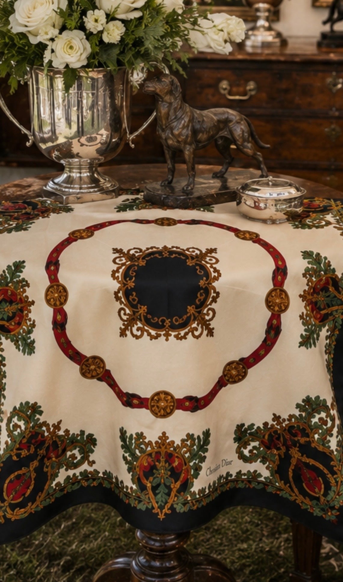 An ornate embroidered tablecloth with a cream base, featuring a large central oval motif in dark green, red, and gold, surrounded by matching floral and scroll patterns. The tablecloth is styled on a dark wooden table with a silver urn of white roses, a bronze horse statue, and a small silver bowl in the background.