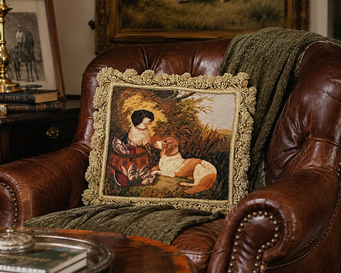 A square decorative throw pillow with a detailed vintage-style tapestry print of a woman and a dog, framed by a textured beige bouclé border, resting on a brown leather armchair with a green throw blanket.