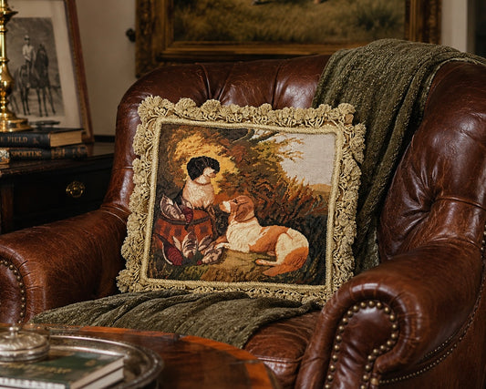 A square decorative throw pillow with a detailed vintage-style tapestry print of a woman and a dog, framed by a textured beige bouclé border, resting on a brown leather armchair with a green throw blanket.