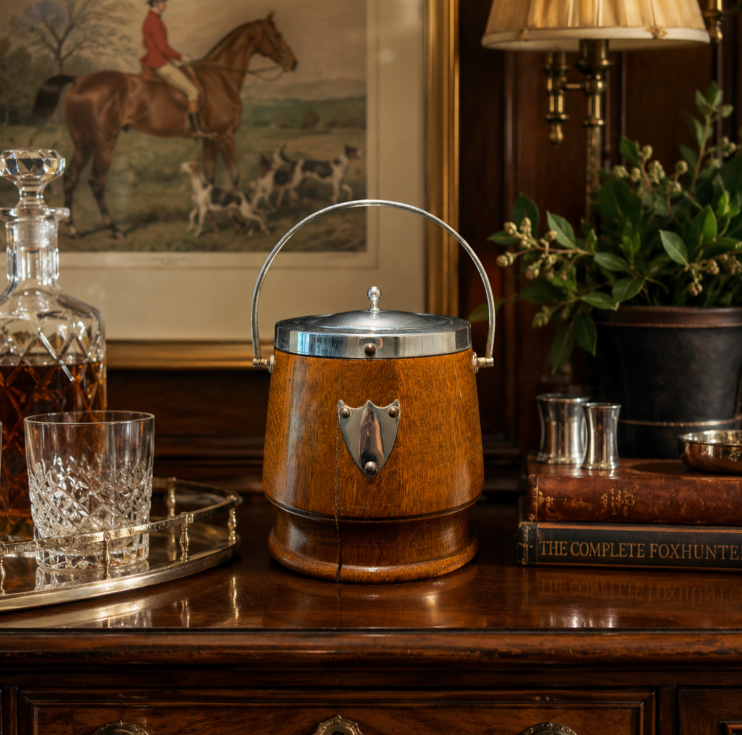 A wooden ice bucket with a silver metal handle and shield-shaped metal detail, displayed on a dark wood table in a styled interior setting with a decanter, glass, books, and framed artwork.