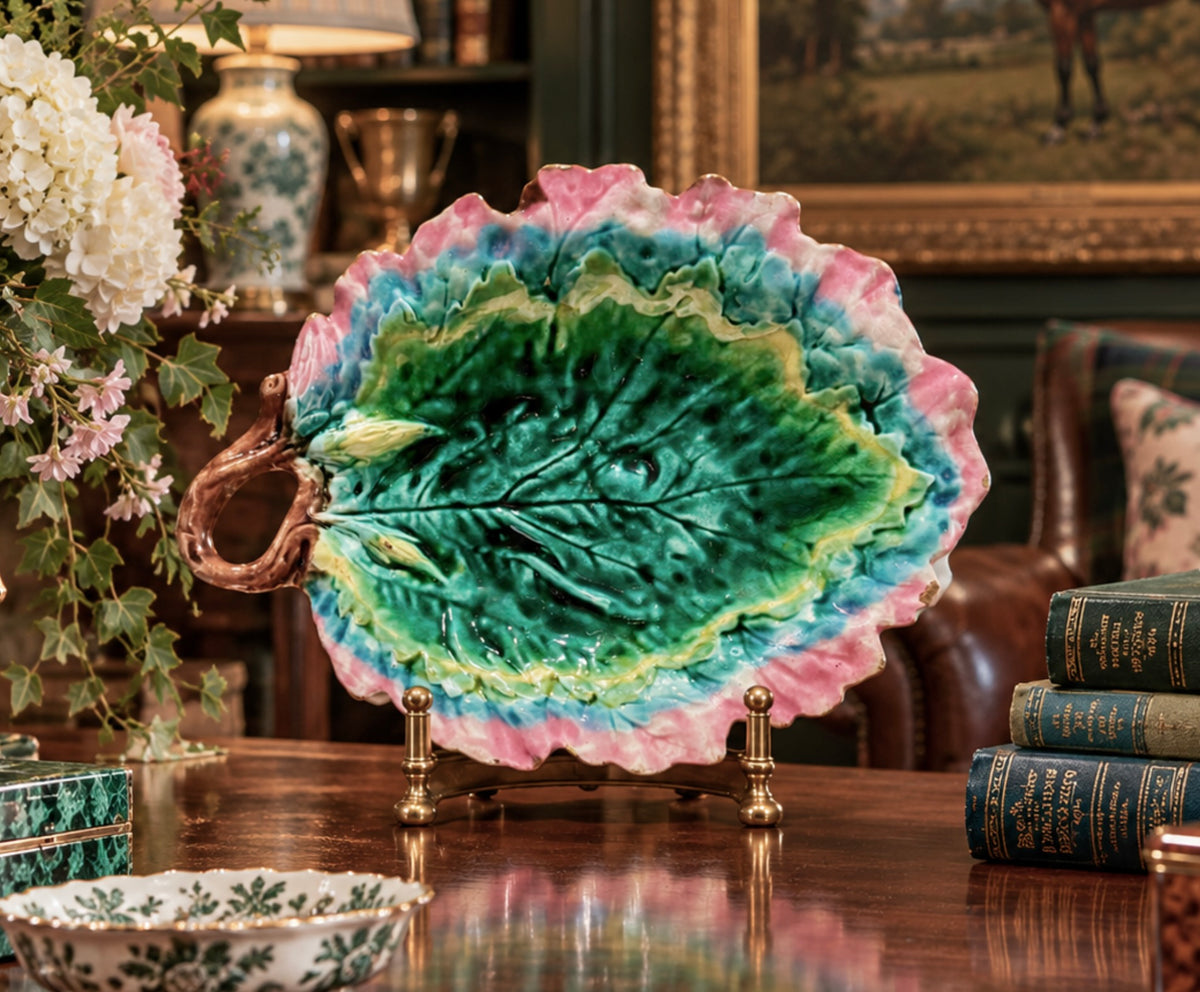 A decorative ceramic leaf-shaped platter with a green center, blue and pink outer edges, and a brown handle, displayed on small brass stands on a polished wooden table in an ornate room.