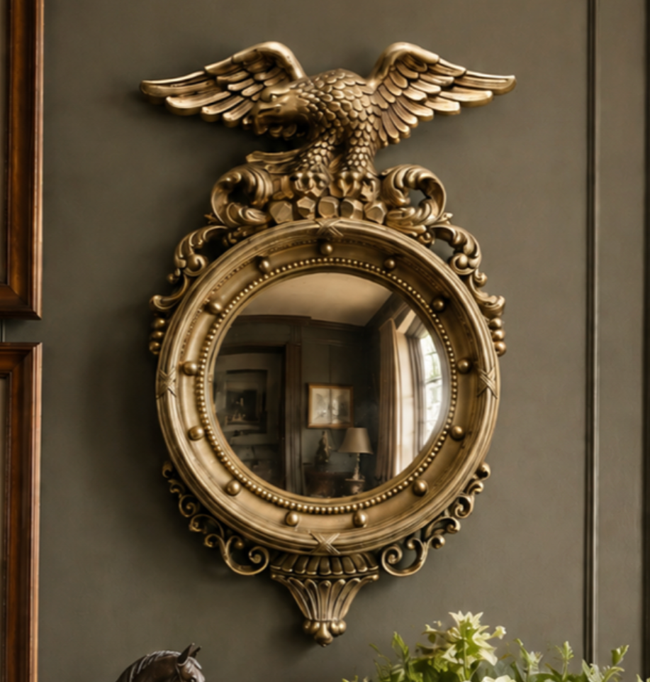 An ornate round wall mirror with an elaborate gold eagle and scrollwork frame, mounted on a dark gray paneled wall in a styled interior setting.