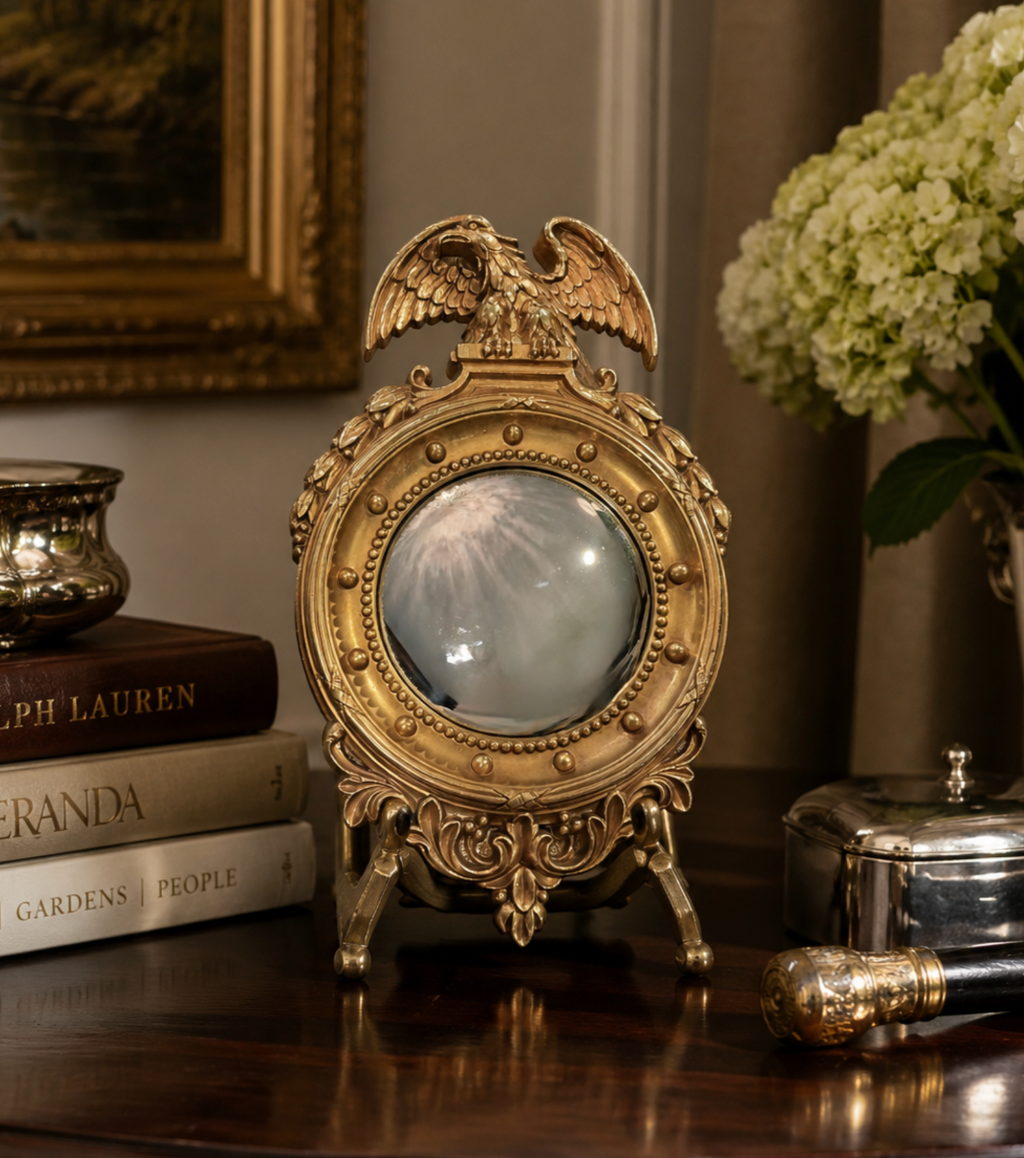 An ornate gold-toned decorative mirror with an eagle crest and round glass center, displayed on a polished wooden table in a styled interior setting with books, flowers, and metal containers.