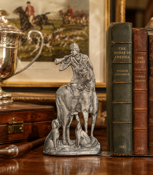 A silver-toned metal figurine of a soldier on horseback blowing a horn, displayed on a polished wooden table next to vintage books.