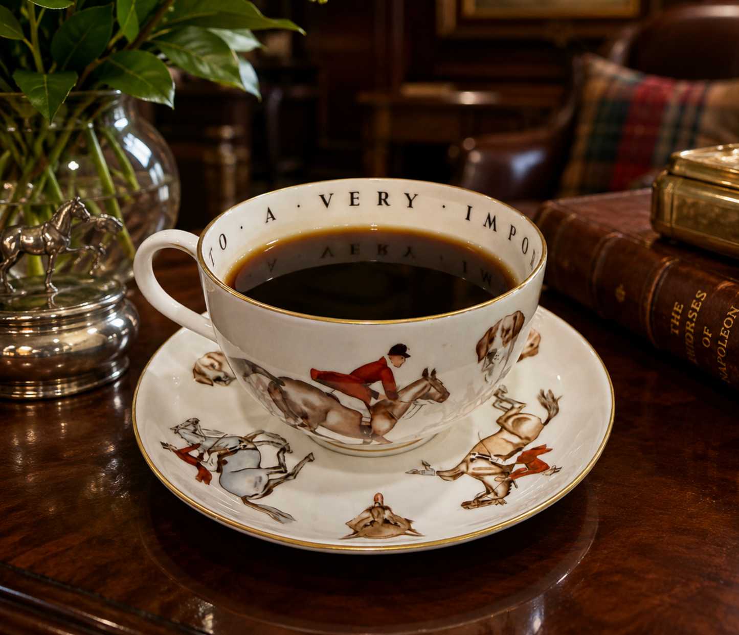 A white porcelain teacup and saucer set filled with black coffee, placed on a dark wooden table in a cozy indoor setting. The cup and saucer are decorated with illustrations of horses and riders and the text 'TO A VERY IMPONDER.