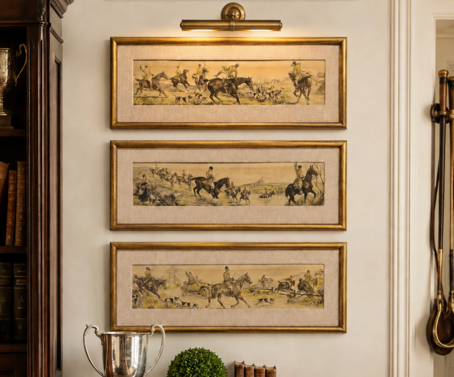 Three framed horizontal art prints depicting equestrian hunting scenes, arranged vertically on a cream-colored wall in a styled interior setting with a gold wall sconce above and decorative objects below.