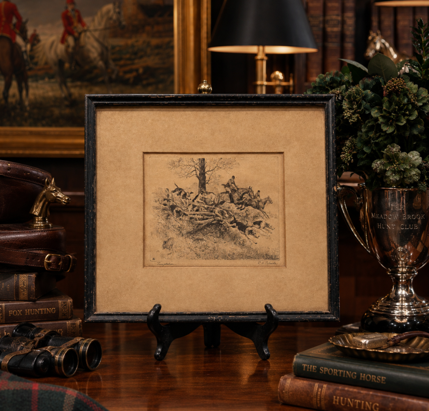 A framed antique-style hunting sketch print displayed on a black easel stand on a wooden table, surrounded by vintage books, a lamp, and a silver trophy cup in a richly decorated room.