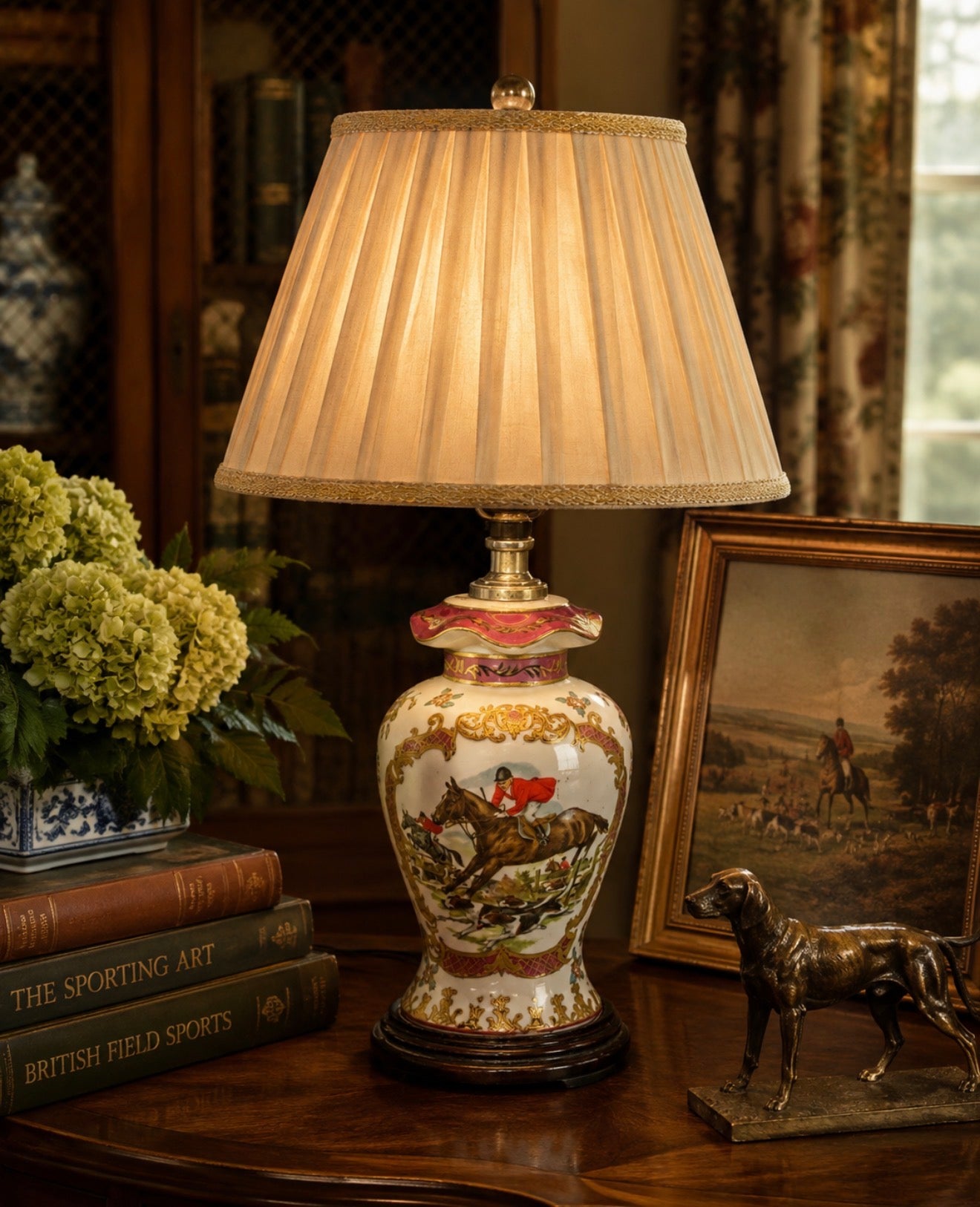 An ornate table lamp with a pleated beige shade and a porcelain-style base decorated with a horse-and-rider scene, styled on a wooden table with books, a framed landscape print, and a dog statue.