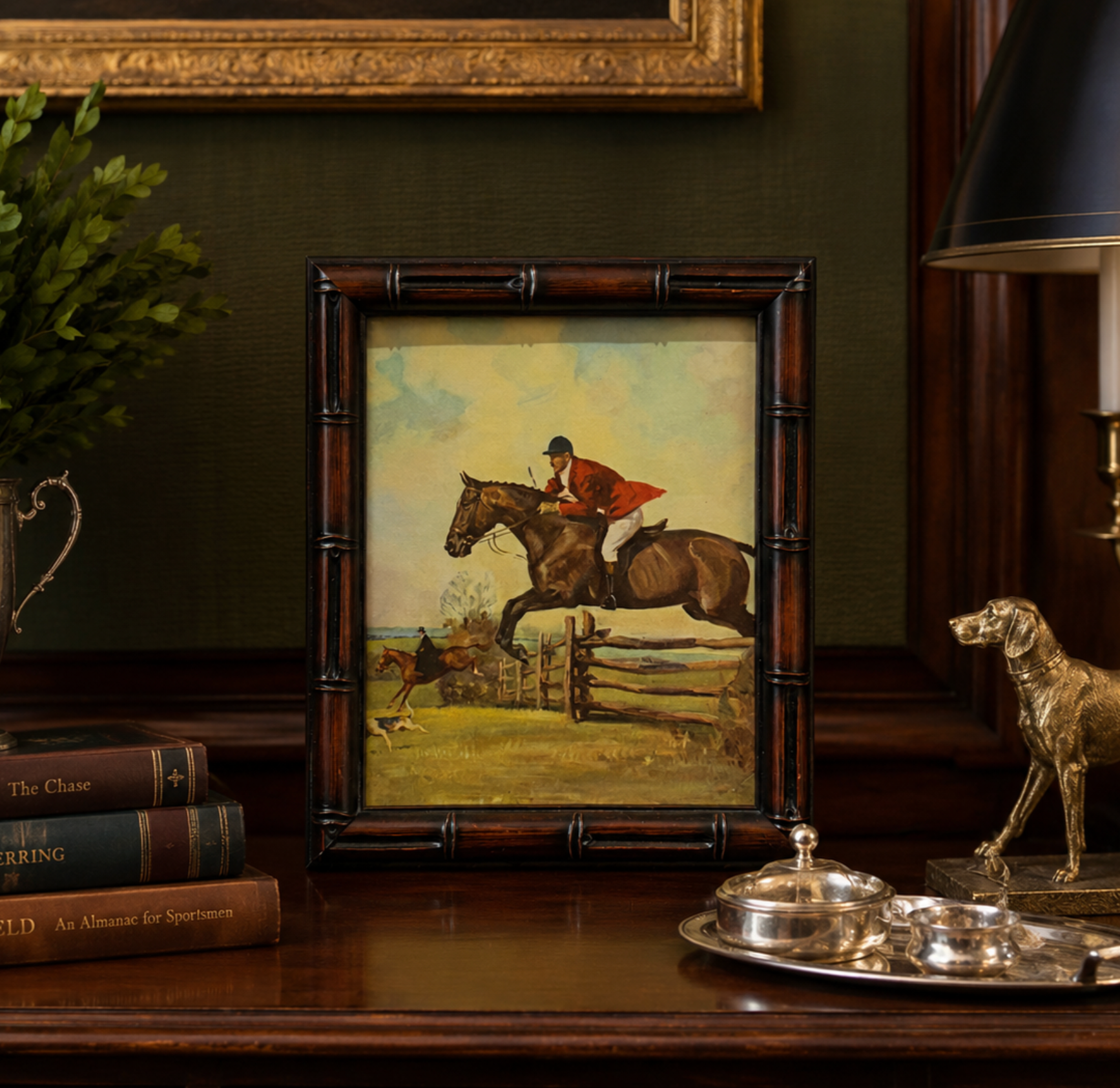 A framed equestrian artwork displayed on a dark wooden table, featuring a rider in red jumping over a horse near a fence. The frame is styled in a classic indoor setting with books, a dog statue, and a lamp.