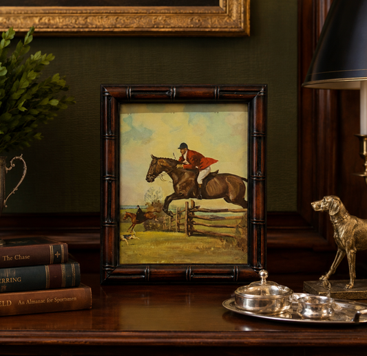 A framed equestrian artwork displayed on a dark wooden table, featuring a rider in red jumping over a horse near a fence. The frame is styled in a classic indoor setting with books, a dog statue, and a lamp.