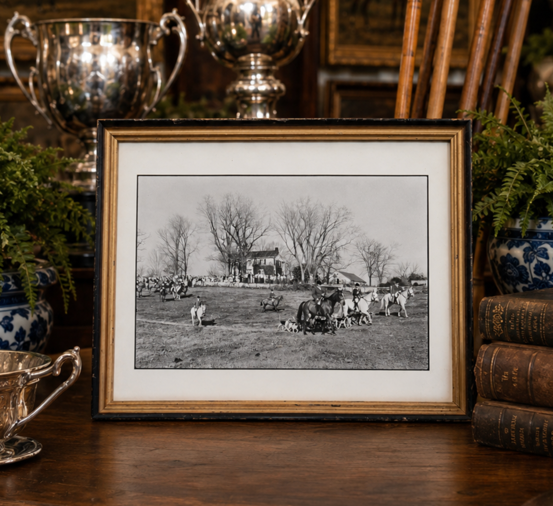 A framed black and white historical photograph print displayed on a wooden table, surrounded by decorative props including silver cups, books, and potted ferns.
