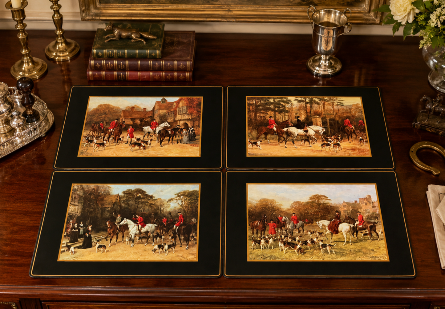 Four framed hunting art prints arranged on a dark wooden table, surrounded by decorative objects like candlesticks, books, and a silver cup.