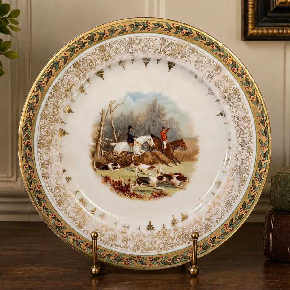 A decorative porcelain hunting plate displayed on a gold stand on a wooden table. The white plate features an ornate gold and green patterned border and a central hand-painted scene of horse hunters with hounds in a wooded setting.