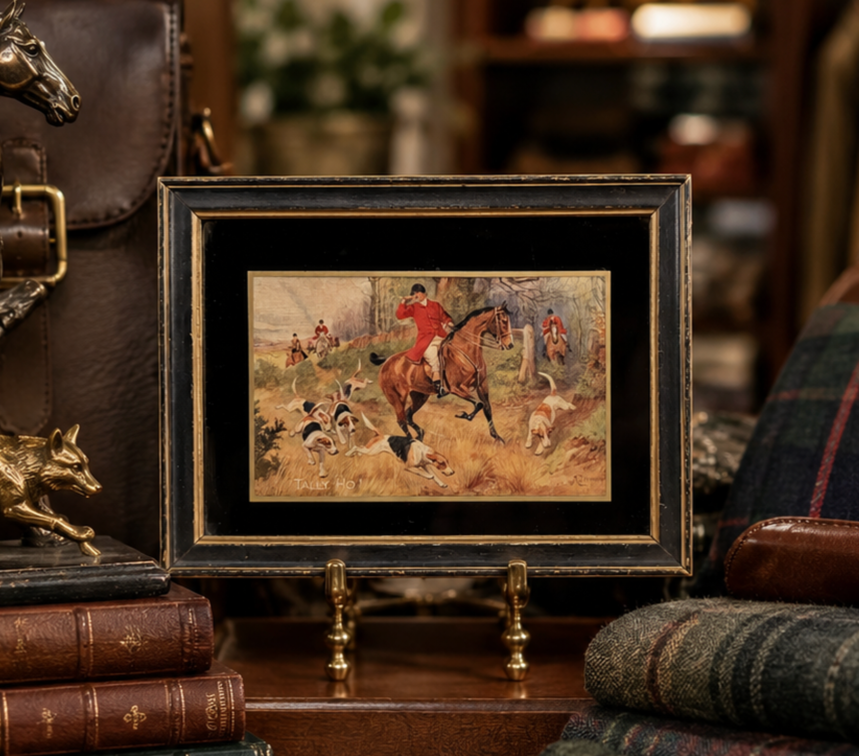 A small framed vintage hunting artwork displayed on a wooden stand in a rustic, dark-themed room. The artwork depicts a horse rider in red on a brown horse, surrounded by dogs, and is set in a black frame with gold trim.