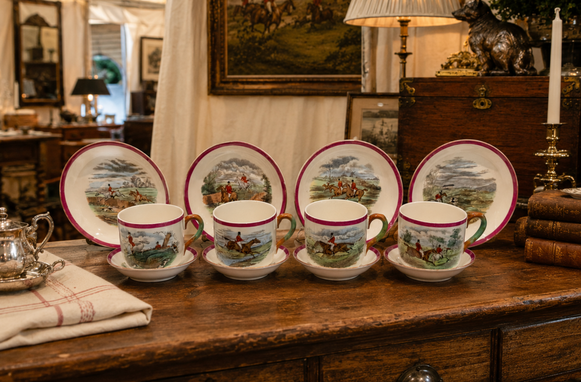 A set of four hand-painted porcelain tea cups with saucers and matching plates, displayed on a rustic wooden chest in a vintage room. The cups and plates feature a red rim and a detailed landscape illustration of a horseback rider on horseback.