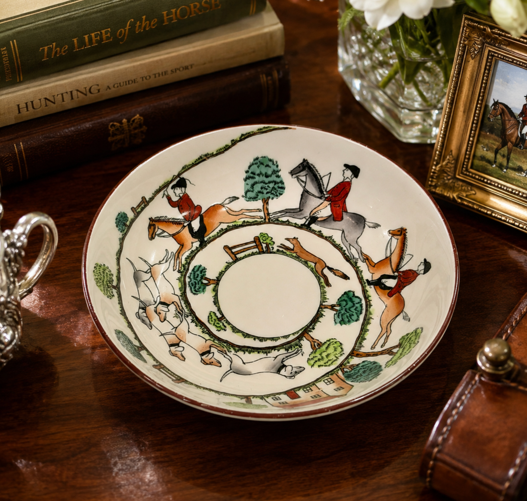 A decorative ceramic plate with a hunting scene illustration, displayed on a dark wooden table surrounded by vintage books, a leather case, and a framed picture.