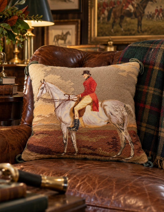 A square woven throw pillow featuring a needlepoint-style depiction of a rider on a horse, styled on a brown leather armchair in a cozy, vintage-filled room.