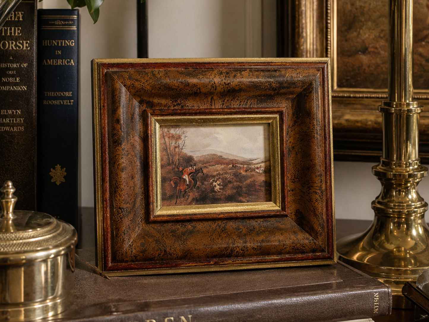 A small framed landscape painting with an ornate dark brown and gold frame, displayed on a shelf next to books and a brass lamp.