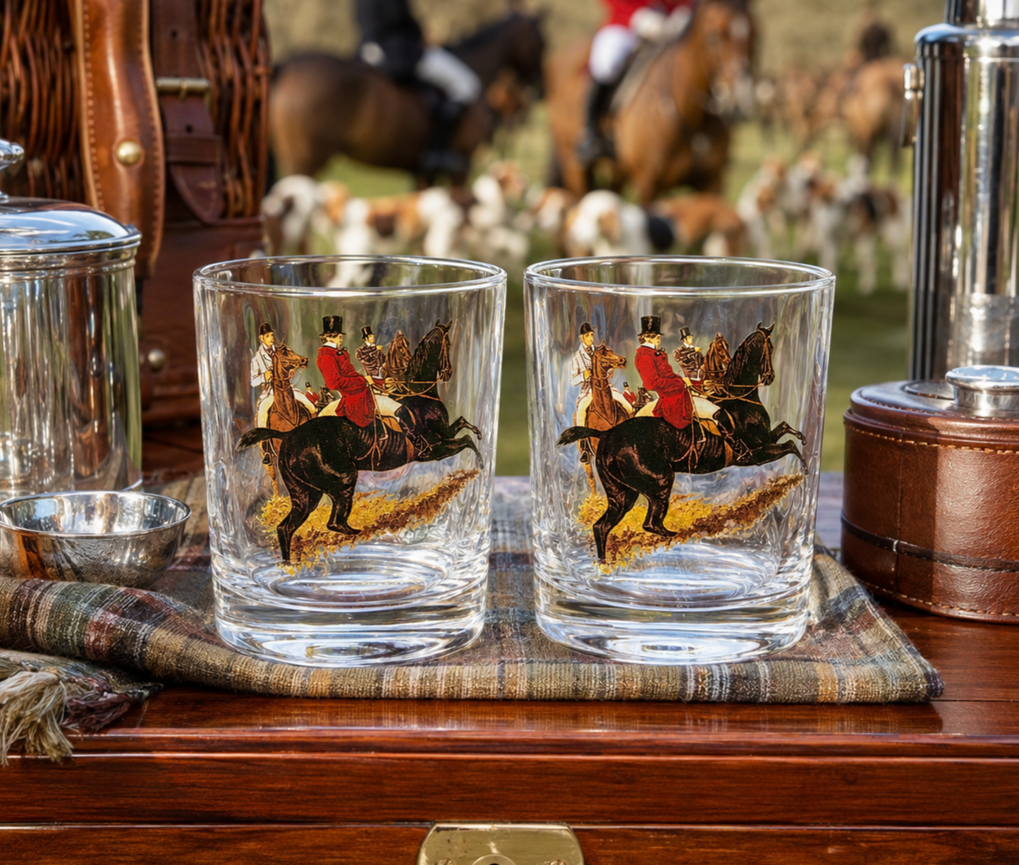 Two clear glass tumblers with a printed hunting scene of riders on horseback jumping a horse, displayed on a wooden surface with a plaid cloth and vintage metal containers around them.