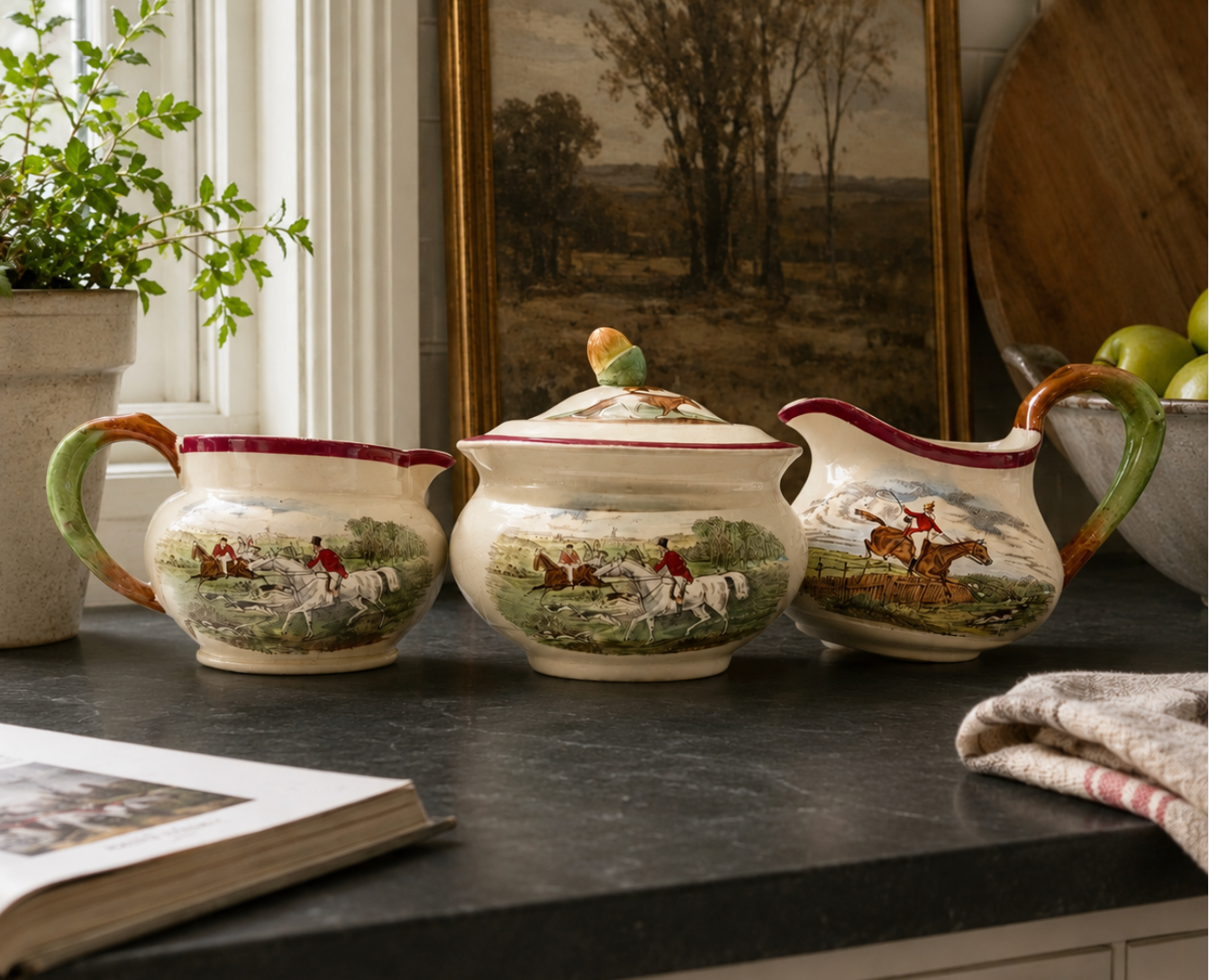 Three ceramic pots with hunting scene designs on a dark surface, with a plant and book in the background.