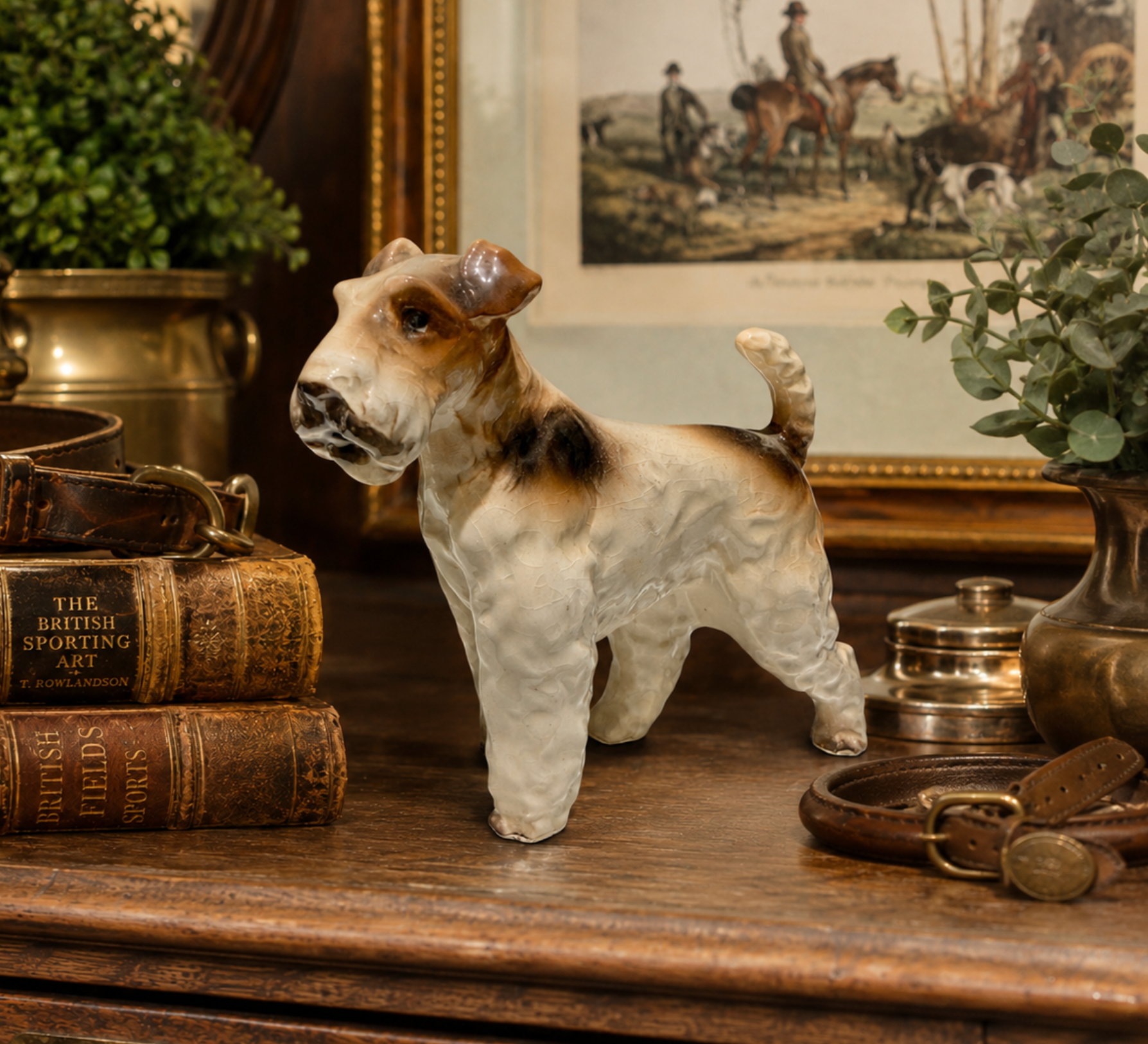 A ceramic figurine of a terrier dog standing on a wooden table, surrounded by vintage books, leather accessories, and potted plants in a styled interior setting.