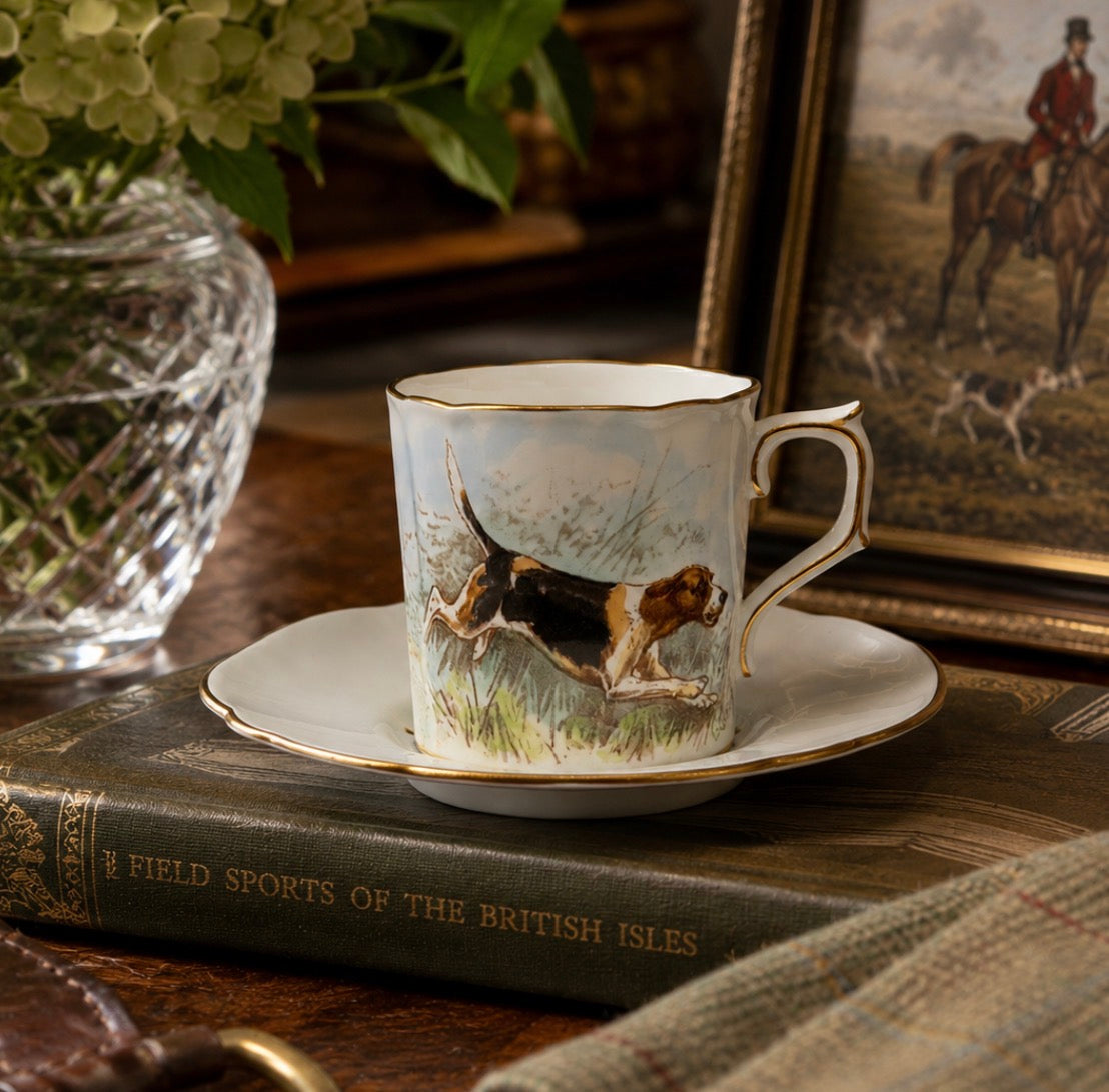A white porcelain teacup and saucer with gold trim, featuring a painted illustration of a hunting dog running through grass, displayed on a vintage book with a glass vase and framed print in the background.
