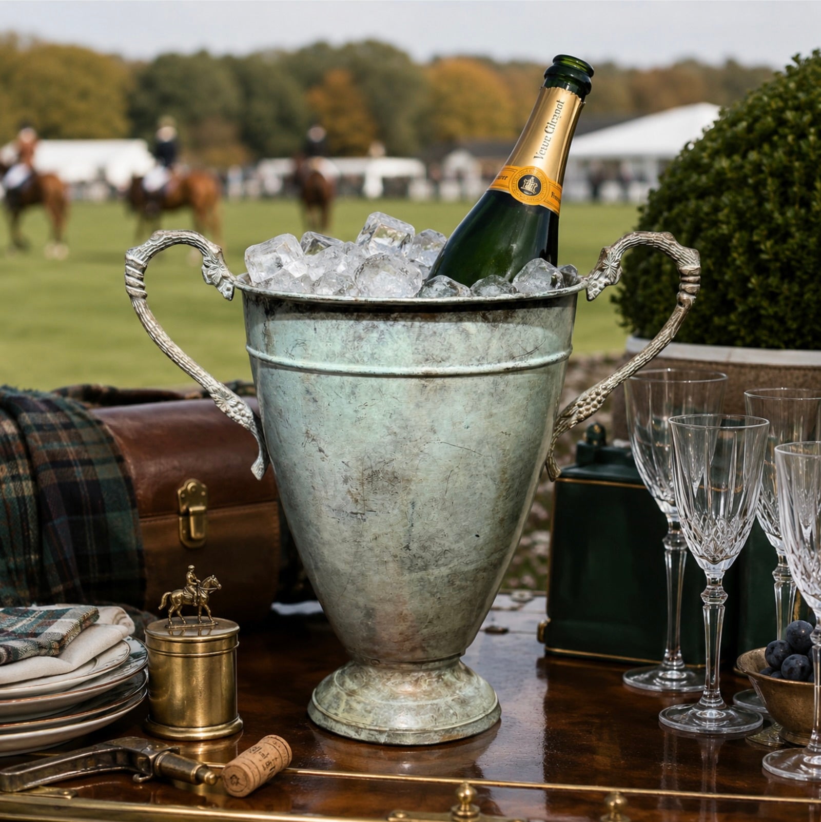 A vintage-style metal champagne bucket filled with ice on a wooden table, with a bottle of champagne leaning inside. Crystal glasses, a small brass canteen, and an ornate horse figurine are arranged around it, with an outdoor polo field and riders blurred in the background.