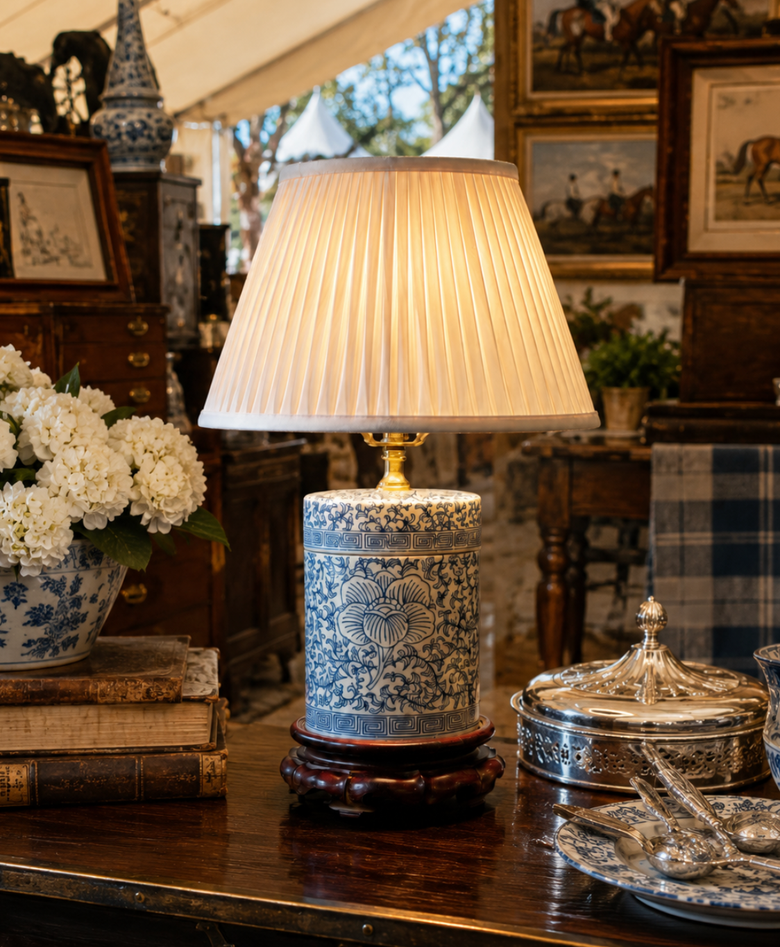 A decorative table lamp with a pleated cream fabric shade and a blue-and-white floral ceramic base on a dark wooden pedestal, displayed in a vintage furniture shop setting.
