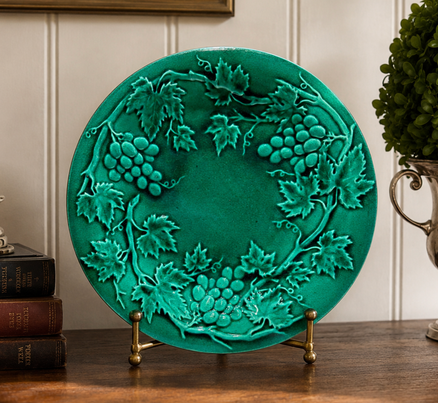 A round teal green decorative plate featuring embossed grapevine and grape motifs, displayed on a small brass stand on a wooden surface.