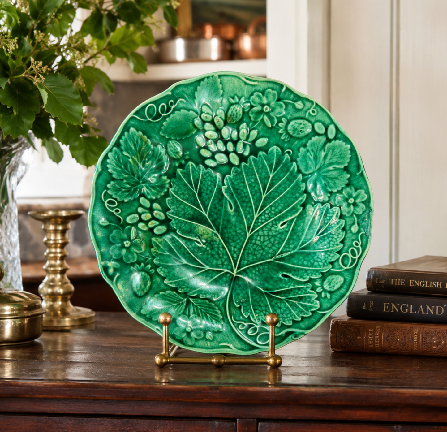 A green glazed ceramic plate with embossed grapevine and leaf motifs, displayed on a small brass stand on a wooden table with books and a vase in the background.