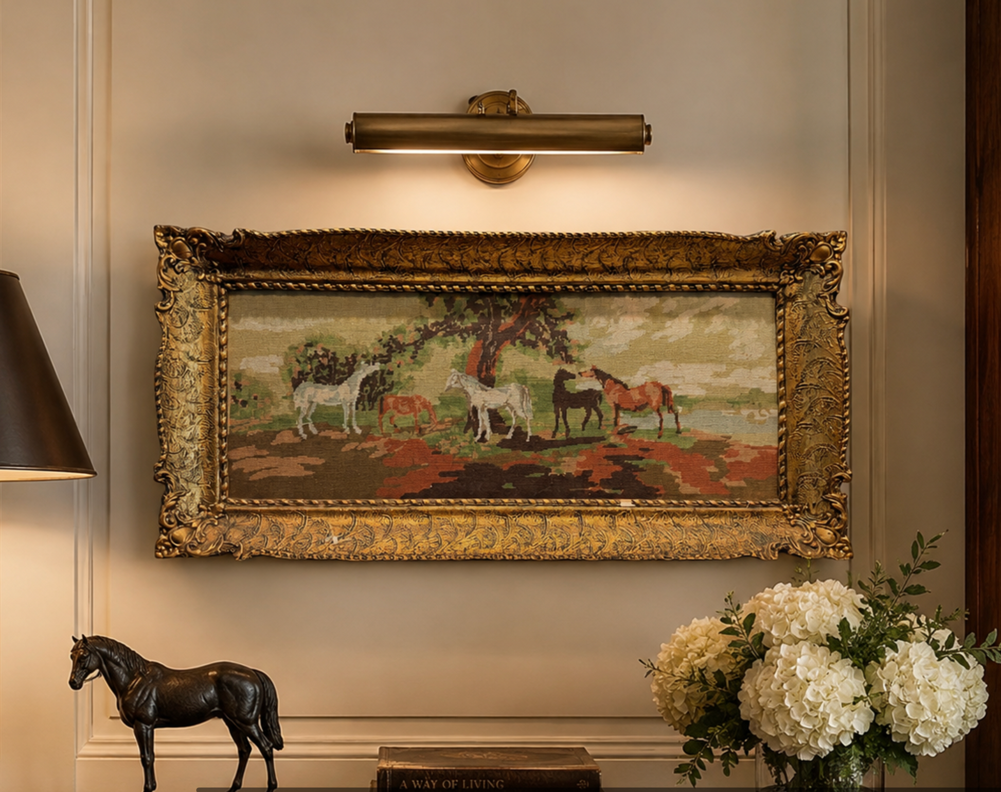 A framed needlepoint tapestry depicting horses in a field under a tree, hung on a beige wall below a brass wall sconce. The frame is ornate gold.